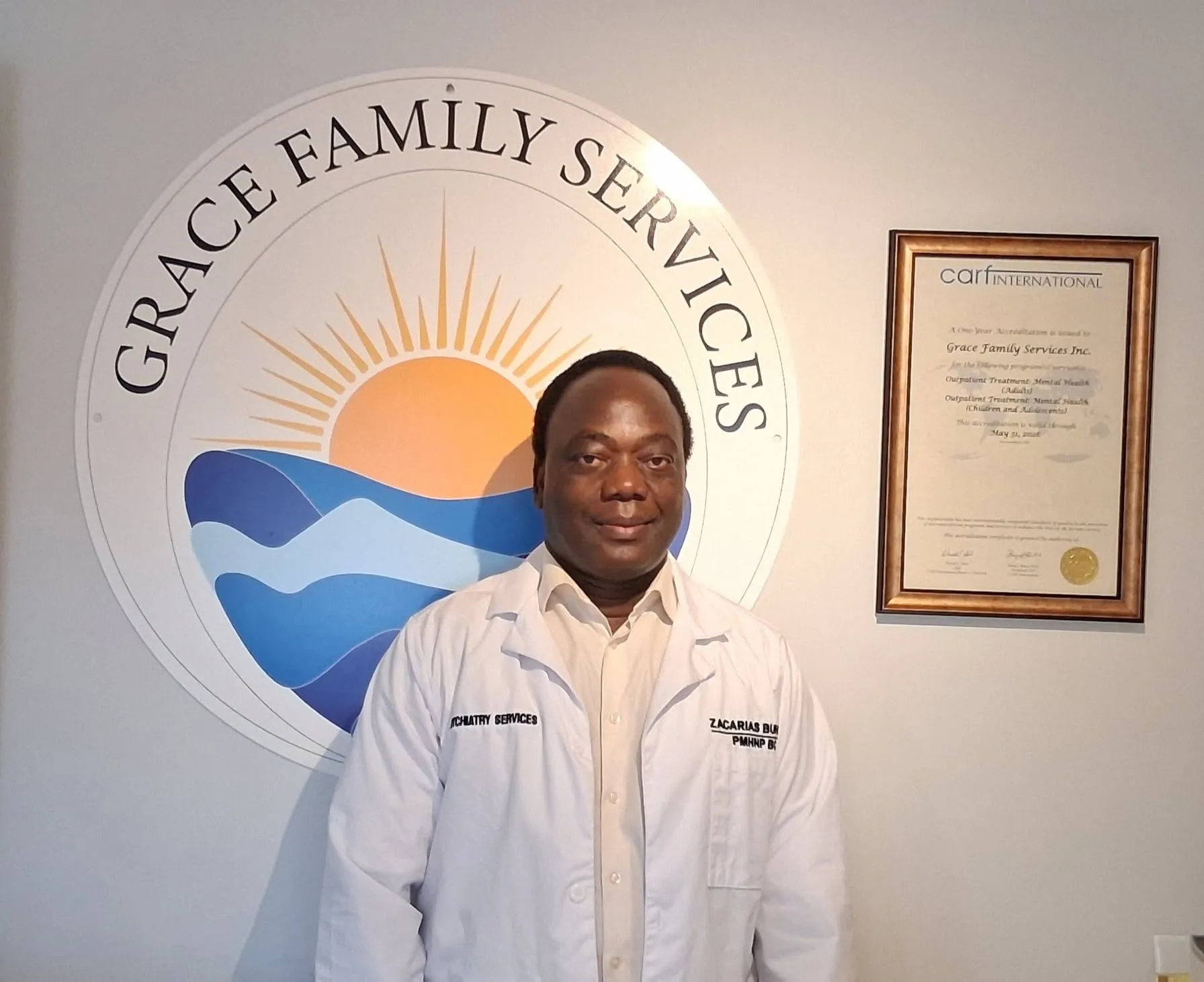Zacarias "Nurse Zac" Buhuro, PMHNP-BC, in a white coat in front of the Grace Family Services logo mural